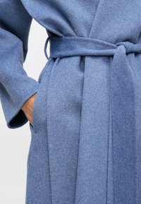 Női kéz puha kék gyapjúkabát zsebében, amelynek derékon megkötött öve van, bemutatva a textúrát és a kényelmes illeszkedést.