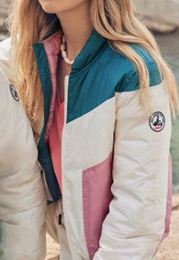 Donna che indossa una giacca color block in teal, bianco e rosa con un patch rotondo JOTT sulla manica e una collana di conchiglie.