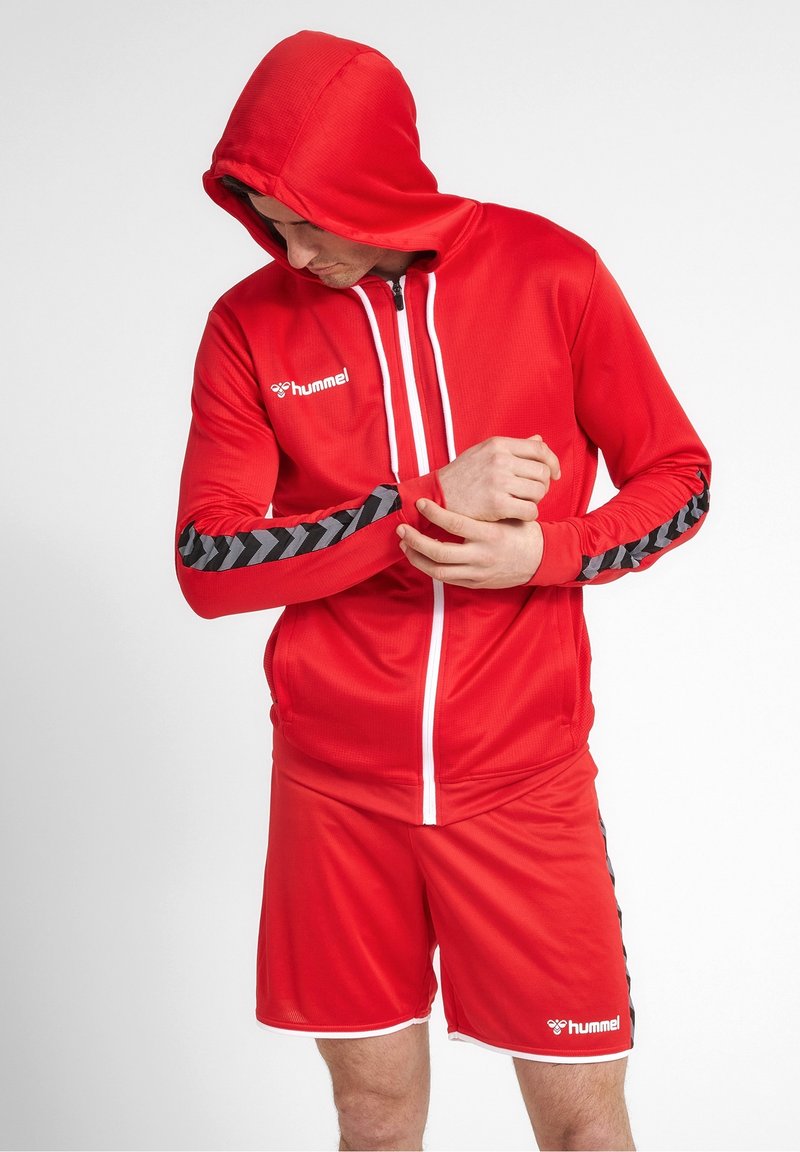 Survêtement de sport rouge comprenant une veste à capuche zippée avec des accents blancs et des rayures noires en chevron sur les manches, assorti d'un short.