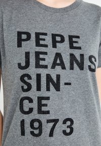 Camiseta de algodón gris con un texto negro en negrita: "PEPE JEANS DESDE 1973" con un patrón texturizado y elevado para mayor énfasis.