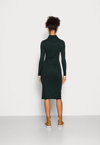 Robe en tricot vert foncé moulante avec col roulé, manches longues et longueur midi, associée à des bottines marron clair. Texture lisse.