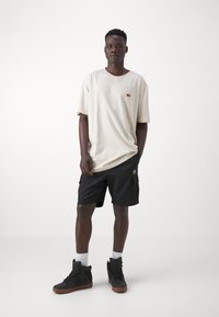 Beige cotton t-shirt with a small embroidered logo, paired with black cargo shorts, and black high-top sneakers. White ankle socks visible.