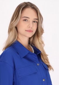 Cămașă cu guler albastru, realizată dintr-un material texturat, care dispune de două buzunare pe piept și închidere cu nasturi în față. Construită cu un finisaj neted și accente metalice.