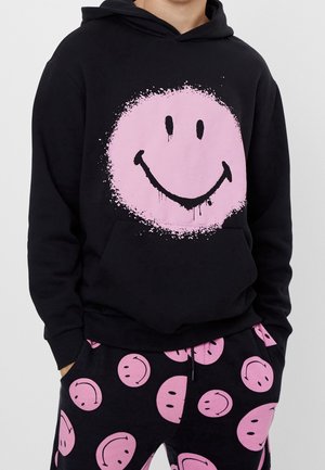 Persona che indossa felpa nera con cappuccio e pantaloni con grandi faccine rosa sorridenti, la felpa presenta una grande faccina sorridente centrata con effetto schizzi di vernice.