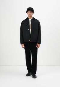 Young man wearing a black button-up jacket, patterned sweater underneath, black trousers, and black sneakers standing against a white background.