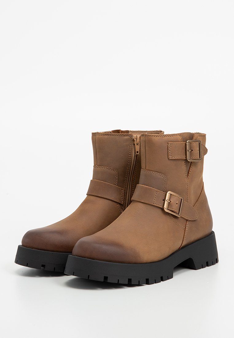 Botas de cuero marrón hasta el tobillo con punta redondeada, hebillas metálicas, cremallera lateral y suela de goma negra gruesa. Acabado texturizado en el cuero.