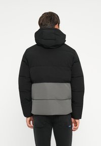 Sort og grå puffervest med hætte, med et syet design, kontrasterende vandrette paneler og en blød, struktureret overflade.