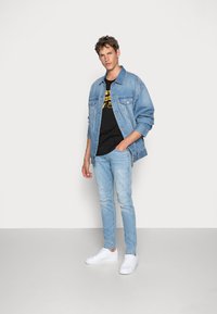 Chaqueta de mezclilla en azul claro con dos bolsillos frontales, llevada sobre una camiseta negra con texto gráfico amarillo, combinada con jeans azules claros y zapatillas blancas.