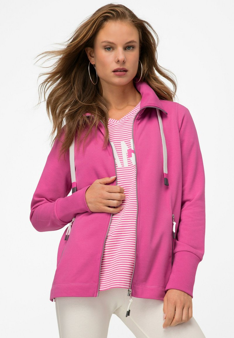Giacca fucsia con zip e cappuccio, dotata di lacci grigi, tasche con zip e polsini a coste. Indossata sopra una maglietta rosa a righe.