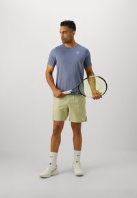 Meeste helesinise lühikese varrukaga spordipluus, millel on logo, koos beeži mustriga lühikesed püksid. Hoides tennisereketit, kantud valgeid treeningjalatseid ja sokke.
