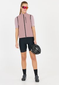 Hellrosa ärmellose Radweste mit einem Reißverschluss vorne, schwarzen Akzenten und einer Tasche. Schwarze Radhosen und einen schwarzen Helm in der Hand.