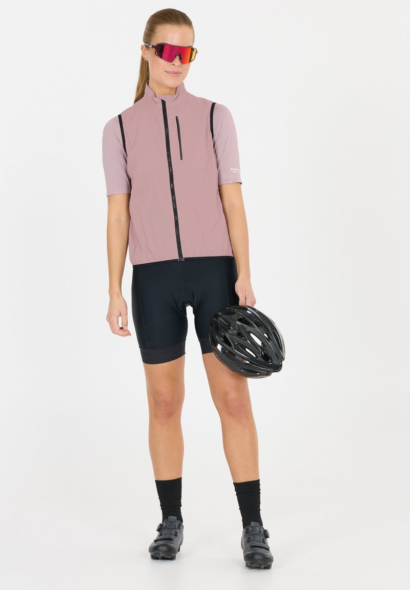 Hellrosa ärmellose Radweste mit einem Reißverschluss vorne, schwarzen Akzenten und einer Tasche. Schwarze Radhosen und einen schwarzen Helm in der Hand.