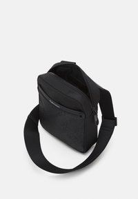 Borsa a tracolla nera con finitura liscia e opaca. Presenta una zip sulla parte superiore, una tracolla regolabile e un design con logo in rilievo su un lato.