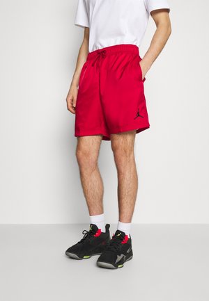 Mężczyzna w czerwonych sportowych szortach z czarnym logo Jordan, białej koszulce, białych skarpetkach i czarnych butach sportowych z czerwonymi i neonowo zielonymi detalami stoi przy białej ścianie.