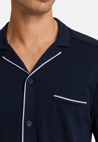 Chemise de pyjama bleu marine avec un col en V, passepoil blanc, deux boutons violets et une poche poitrine avec une ligne d'accent blanc.