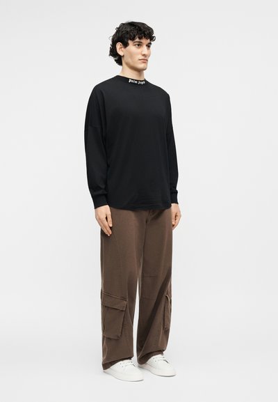 Giovane uomo in piedi che indossa una maglia nera a maniche lunghe con colletto "Palm Angels", pantaloni cargo marroni e sneakers bianche su sfondo neutro.