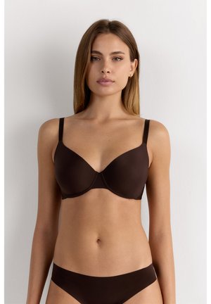 Femme aux cheveux raides portant un ensemble assorti de soutien-gorge et de sous-vêtements marron foncé, face à un fond clair uni.