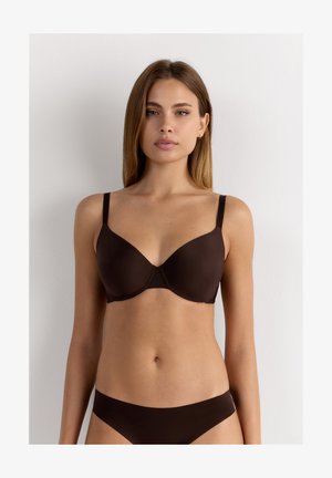Femme aux cheveux raides portant un ensemble assorti de soutien-gorge et de sous-vêtements marron foncé, face à un fond clair uni.