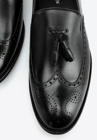 Mocasines de piel negra con borlas y detalles de brogue en la punta y los lados, sobre un fondo blanco.