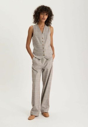 Femme debout, les mains dans les poches, portant un gilet à carreaux gris avec des boutons et un pantalon large assorti, chaussures beige.