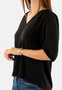 Top nero a V con una vestibilità rilassata e maniche corte arrotolate, caratterizzato da una leggera texture scintillante. Indossato con jeans blu e un bracciale argentato.