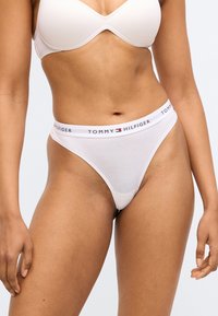 Fehér pamut tangában kontrasztos, márkázott elasztikus derékpánttal. Simaszövetű, minimális takarást biztosító kialakítással.