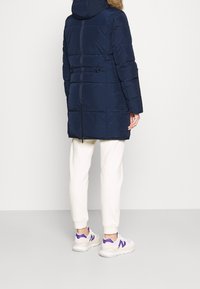 Giacca puffer blu navy con cappuccio foderato in pelliccia, cuciture orizzontali, lati con zip e vestibilità comoda, abbinata a pantaloni della tuta crema e sneakers bianche.