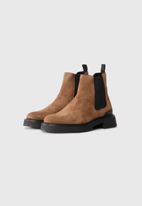 Par de botas de tobillo de ante marrón con paneles laterales elásticos negros y suelas de goma negra gruesas sobre un fondo blanco.
