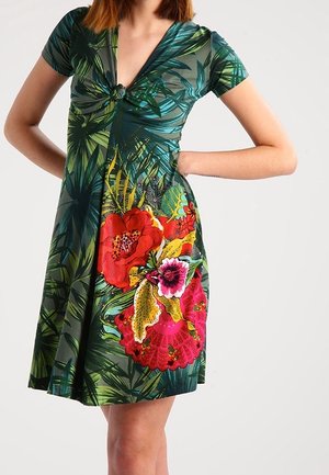 Femme portant une robe courte à manches courtes, longueur genou, avec des motifs de feuilles tropicales vertes et de grands dessins floraux rouges et roses.