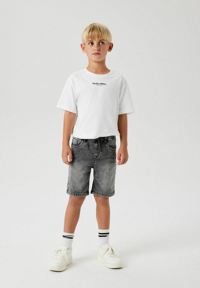 T-shirt en coton blanc avec un texte "édition studio" en noir, associé à un short en denim gris délavé et des baskets blanches avec des rayures noires sur les chaussettes.