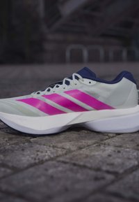 Chaussure de course blanche et bleu marine avec trois bandes roses sur le côté, posée sur une surface pavée.