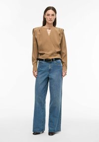 Blouse enveloppante marron avec épaules plissées, associée à un jean en denim bleu ample et une ceinture noire. Le mannequin se tient devant un fond blanc.