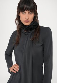 Schwarze Bluse mit langen Ärmeln und Krawatten detail am Hals. Glatte Textur, taillierte Passform. Das Model hat schulterlanges Haar.