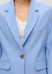 Blazer bleu clair en tissu texturé, avec un revers à cran, un bouton marron unique et deux poches avant.