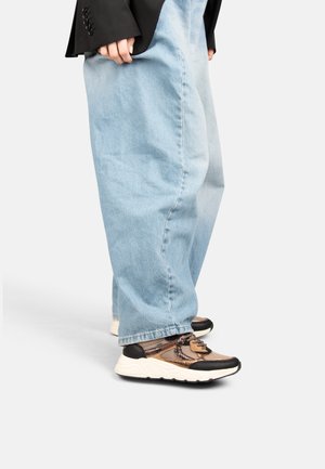 Persona che indossa jeans azzurri a gamba larga e sneakers marroni, nere e bianche con fantasia, in piedi su un pavimento bianco.