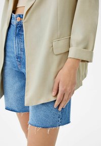 Beige blazer met opgerolde mouwen, gecombineerd met blauwe denim shorts met een rafelige zoom. Focus op de textuur en het rechte ontwerp van beide stukken.