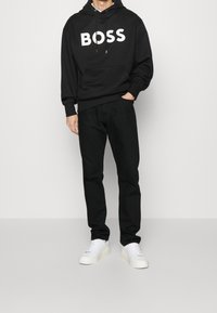 Black hoodie with large white "BOSS" logo, paired with black pants and white sneakers. The fabric appears smooth with a relaxed fit.
