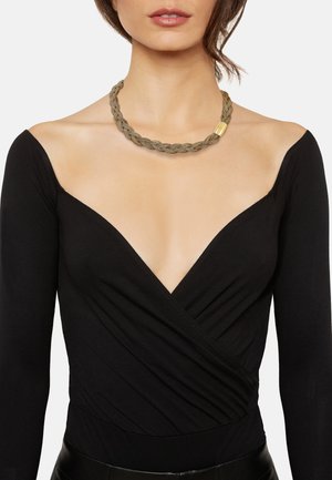 Kvinde iført sort off-shoulder V-hals top og flettet guld halskæde med lås, vist fra hals til talje på hvid baggrund.