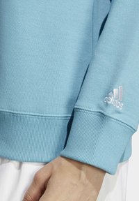 Ljusblå sweatshirtärm med ribbad manschett, vitt Adidas-logga broderad nära handleden, och en del av en hand och vitt plagg synligt nedanför.