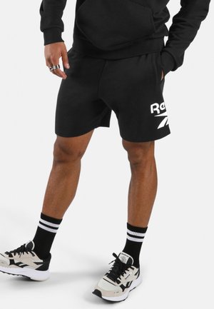 Man die een zwarte hoodie en zwarte Reebok short draagt, zwarte sokken met witte strepen en witte Reebok sneakers met zwarte veters.