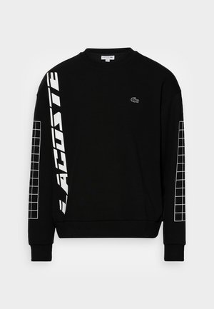 Sweat-shirt noir à manches longues avec des motifs en grille blanche sur les manches, grand texte vertical "LACOSTE" sur le bras gauche et petit logo crocodile sur la poitrine.