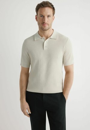 Mężczyzna z krótkimi brązowymi włosami, ubrany w jasnobeżową, prążkowaną koszulkę polo i czarne spodnie, stojący na tle jednolitej białej ściany.