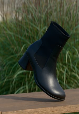 Schwarzer Leder-Knöchelstiefel mit Blockabsatz, stehend auf einer Holzoberfläche mit grünen Pflanzen im Hintergrund.