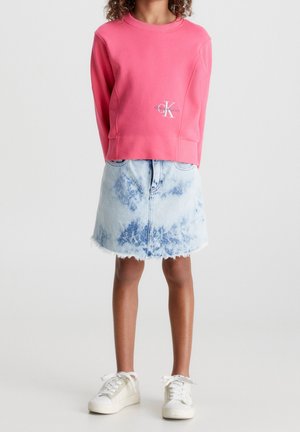 Niña con una camiseta de manga larga rosa con un logo, falda de mezclilla azul claro efecto ácido y zapatillas blancas, de pie con los brazos detrás de la espalda.