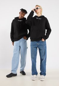Felpe nere con la scritta "New York", abbinate a jeans larghi blu chiaro e blu scuro. Un modello indossa occhiali da sole e sneaker.