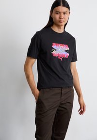 Black cotton t-shirt featuring colorful "PUMA Forever Legends" text design across the chest, paired with brown trousers.