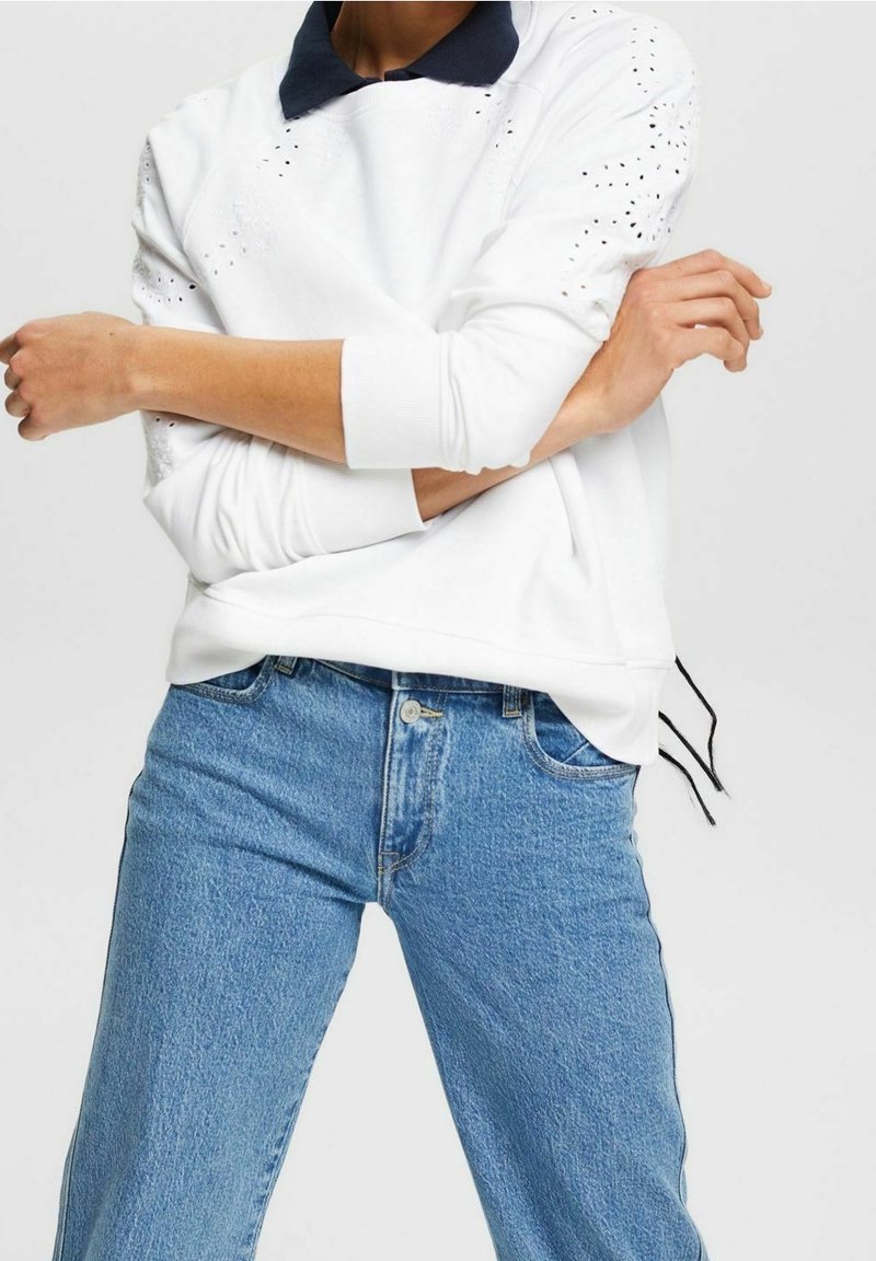 Personne portant un sweat-shirt blanc avec des détails perforés et un jean bleu, les bras croisés à la taille.