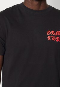 Černé bavlněné tričko s krátkými rukávy a kulatým výstřihem, na přední straně s výrazným červeným textem v gotickém písmu.