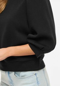 Femme portant un haut noir à manches longues bouffantes retroussées et un jean en denim bleu clair sur un fond blanc.