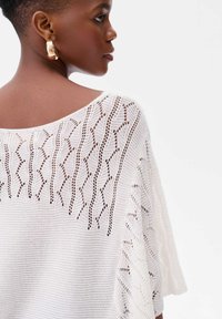 Witte gebreide poncho met een losse, open geweven ontwerp en een golvend patroon langs de schouders, afgewerkt met een zachte, textuur. 